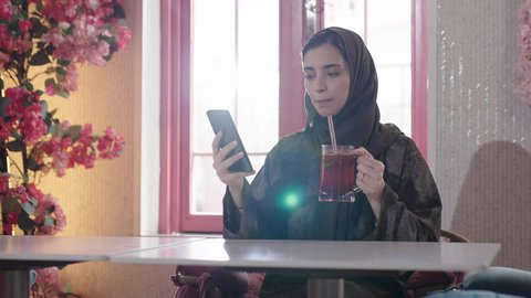 قضاء الأوقات الممتعة في الخارج، الاستمتاع بالمشروبات الباردة في فصل الصيف, الاستمتاع بالأنشطة الترفيهية, امرأة عربية خليجية سعودية محجبة ترتدي عباءة تجلس في كافي شوب راقي تقوم باستخدام الهاتف الجوال، إيماءات السعادة و السرور