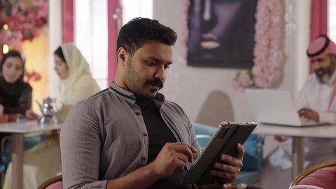 إدارة شؤون الأعمال وتنظيمها عبر الاجهزة التقنية، لقاء الاصدقاء في أوقات الفراغ، أجواء الهدوء والاسترخاء داخل كافيه راقي، تصوير مقرب لرجل خليجي عربي سعودي يرتدي الزي الكاجوال يجلس على الكرسي يعمل على جهاز التابلت اللوحي، قضاء أوقات ممتعة في الويكند