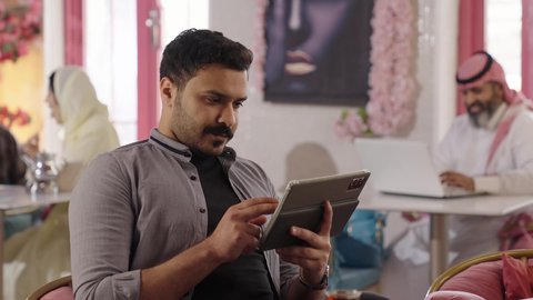 تنظيم وإدارة شؤون الأعمال عبر الاجهزة التقنية، لقاء الاصدقاء في أوقات الفراغ، أجواء الهدوء والاسترخاء داخل كافيه راقي، تصوير مقرب لرجل خليجي عربي سعودي يرتدي الزي الكاجوال يجلس على الكرسي يعمل على جهاز التابلت اللوحي، قضاء أوقات ممتعة في الويكند