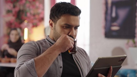 تعابير السرور والراحة، تنظيم وإدارة شؤون الأعمال عبر الاجهزة التقنية، لقاء الاصدقاء في أوقات الفراغ، أجواء الهدوء والاسترخاء داخل كافيه راقي، تصوير مقرب لرجل خليجي عربي سعودي يرتدي الزي الكاجوال يجلس على الكرسي يعمل على جهاز التابلت اللوحي، قضاء أوقات ممتعة في الويكند