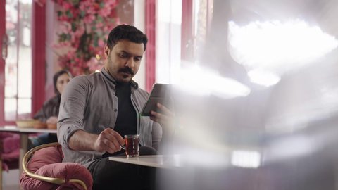 التواصل مع الأهل والأصدقاء، دمج العمل مع التقنية والتكنولوجيا، أجواء الهدوء والاسترخاء داخل كافيه راقي، تصوير مقرب لرجل خليجي عربي سعودي يرتدي الزي الكاجوال يجلس على الكرسي يعمل على جهاز التابلت اللوحي ويشرب الشاي، قضاء أوقات ممتعة في الويكند