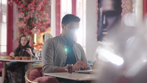 العمل في احدى كافيهات ومطاعم، دمج العمل مع التقنية والتكنولوجيا، أجواء الهدوء والاسترخاء داخل كافيه راقي، تصوير مقرب لرجل خليجي عربي سعودي يرتدي الزي الكاجوال يجلس على الكرسي يعمل على جهاز احاسب المحمول ويشرب الشاي، قضاء أوقات ممتعة في الويكند