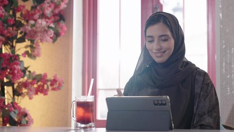 Success and achieving goals, developing businesses and strategies, communicating with family and friends, using a laptop, a veiled Saudi Arabian woman wearing an abaya sits on a sofa in a high-end cafe, enjoying moments of peace and relaxation and drinking cold juices.