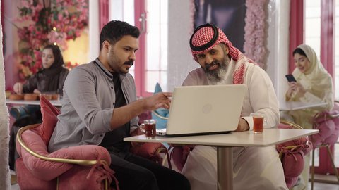 DialogueDialogue and discussion among colleagues using a laptop to complete projects and tasks in a cafe spending enjoyable time with friends a close-up photo of a Saudi Arabian man wearing a thobe and shemagh sitting with his friend in a cafe drinking red tea in a cafeintegrating technology into daily life..