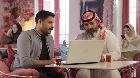 Use your laptop to complete projects and tasks in the cafe, chat and discuss with colleagues, spend enjoyable time with friends. A close-up photo of a Saudi Arabian man wearing a thobe and shemagh sitting with his friend in a cafe, drinking red tea. Integrating technology into daily life.
