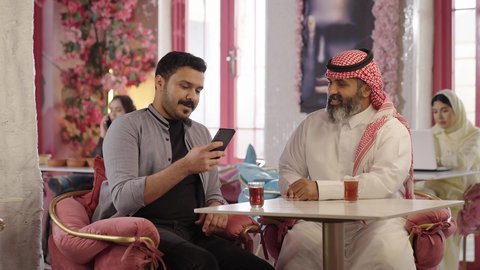 توثيق اللحظات السعيدة مع الصديق في الكافيه، الحوار والنقاش بين زملاء العمل، قضاء وقت ممتع مع الأصدقاء, صورة مقربة لرجل عربي خليجي سعودي يرتدي الثوب و الشماغ يجلس مع صديقه في المقهى، شرب الشاي الاحمر في كافيه، دمج التكنولوجيا في الحياة اليومية