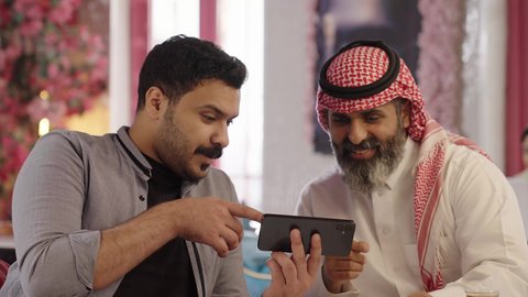Watching a match on a mobile phone screen, conversations and discussions among colleagues, spending quality time with friends, a close-up photo of a Saudi Arabian man wearing a thobe and shemagh sitting with his friend in a cafe, drinking red tea in a cafe, integrating technology into daily life