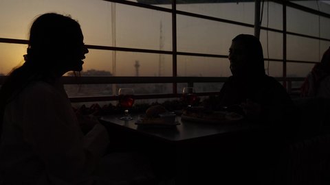 جلسة وناسة يوم الاجازة، وقت الغروب التصوير في الظل، تعابير السعادة والسرور بلمة الرفاق، كوفي شوب راقي في المملكة، الاستمتاع بالمشروبات الباردة، تطوير العلاقات الإجتماعية، التسامر والدردشة مع الأصدقاء، امرأة عربية خليجية سعودية محجبة تجلس مع صديقتها في المطعم