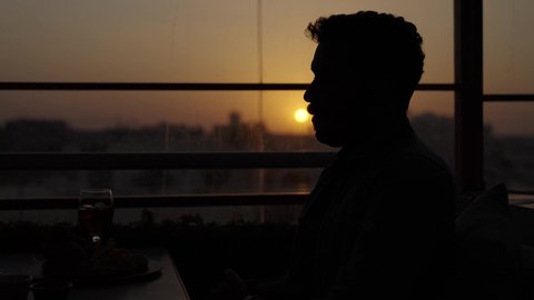 Expressing love and affection between friends, a sunset photoshoot in the shade, working in a cafe or restaurant, the calm and relaxing atmosphere inside a classy cafe, a close-up of a Gulf Arab Saudi man in casual attire sitting in a chair holding a glass of cold drinks, enjoying a pleasant weekend.