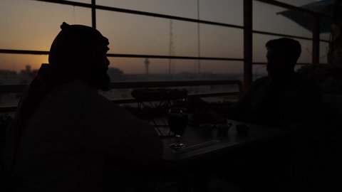 تصوير في وقت الغروب، الحوار والنقاش بين زملاء العمل، قضاء وقت ممتع مع الأصدقاء, صورة مقربة لرجل عربي خليجي سعودي يرتدي الثوب و الشماغ يجلس مع صديقه في المقهى، شرب الشاي الاحمر في كافيه، دمج التكنولوجيا في الحياة اليومية
