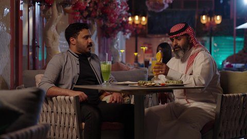 Chatting with friends, enjoying weekends, sipping drinks, a close-up of a Saudi Arabian man wearing a thobe and shemagh sitting in a cafe with his friend, gestures of happiness and joy, eating sweets and pastries at a classy cafe.