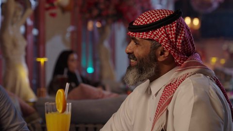 الاستمتاع بشرب المشروبات الباردة، لقاء الاصدقاء في أوقات الفراغ، أجواء الهدوء والاسترخاء داخل كافيه راقي، تصوير مقرب لرجل خليجي عربي سعودي يرتدي الثوب التقليدي والشماغ يجلس على الكرسي يحمل كأس من عصير البرتقال الطبيعي، قضاء أوقات ممتعة في الويكند
