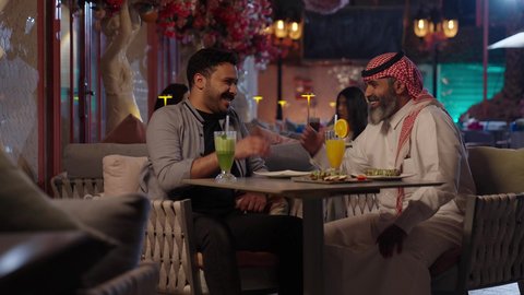 ضربة يد دليل توافق وجهات النظر بين الاصحاب، قضاء أوقات ممتعة في الويكند، الاستمتاع بتناول المشروبات ، تصوير مقرب لرجل عربي خليجي سعودي يرتدي الثوب و الشماغ يجلس فى المقهي برفقة صديقة، ايماءات السعادة و السرور، تناول الحلويات والمخبوزات الجاهزة كافيه راقي