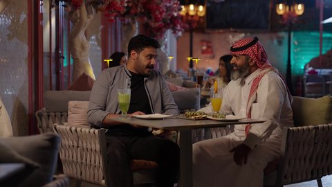 Taking a selfie with friends, enjoying a fun weekend, sipping drinks, a close-up of a Saudi Arabian man wearing a thobe and shemagh sitting in a cafe with his friend, gestures of happiness and joy, eating sweets and ready-made pastries, a classy cafe