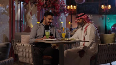 A selfie with friends, enjoying a fun weekend, sipping drinks, a close-up of a Saudi Arabian man wearing a thobe and shemagh sitting in a cafe with his friend, gestures of happiness and joy, eating sweets and pastries at a classy cafe.