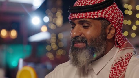 The concept of prestige and elegance, meeting friends during leisure time, the atmosphere of tranquility and relaxation inside a sophisticated cafe, a close-up photograph of a Gulf Arab Saudi man wearing a traditional thobe and shemagh, sitting in a chair holding a glass of fresh orange juice, spending enjoyable times on the weekend.