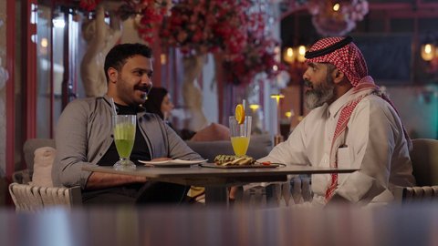 Chatting, socializing, and spending enjoyable weekends having fun with drinks a close-up photo of a Saudi Arabian Gulf man wearing a thobe and shemagh sitting in a cafe with his friend gestures of happiness and joy eating sweets and ready-made pastries a classy cafe.