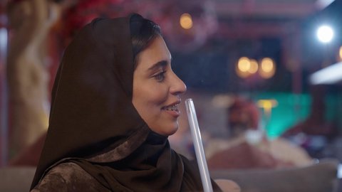 تبادل الاحاديث و الدردشة مع احد الاصدقاء، تصوير مقرب لإمرأة عربية خليجية سعودية ترتدي الحجاب والعباءة تقوم بتدخين الشيشة في إحدى الكافيهات, قضاء أوقات ممتعه في الخارج في عطلة نهاية الاسبوع، جلسة وناسة ونقاشات وحوارات، تدخين الشيشة وتناول المشروبات الصيفية الباردة