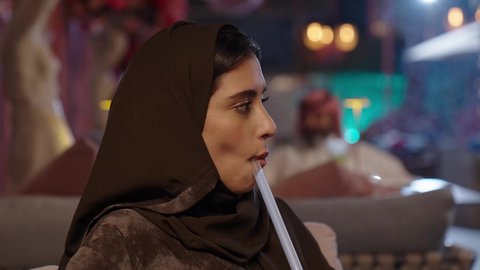 في عطلة نهاية الاسبوع قضاء أوقات ممتعه في الخارج، تبادل الاحاديث و الدردشة مع احد الاصدقاء، تصوير مقرب لإمرأة عربية خليجية سعودية ترتدي الحجاب والعباءة تقوم بتدخين الشيشة في إحدى الكافيهات, جلسة وناسة ونقاشات وحوارات، تدخين الشيشة وتناول المشروبات الصيفية الباردة