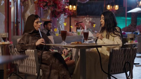 كوفي شوب راقي في المملكة، الاستمتاع بالمشروبات الباردة، جلسة وناسة يوم الاجازة، تطوير العلاقات الإجتماعية، التسامر والدردشة مع الأصدقاء، تعابير السعادة والسرور بلمة الرفاق، امرأة عربية خليجية سعودية محجبة تجلس مع صديقتها في المطعم، صديقتان يقومان بتدخين الشيشة