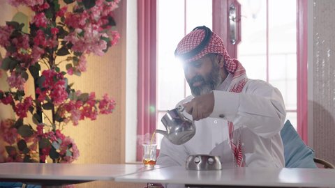 سكب الشاي الاحمر في كأس زجاجي، لقاء الاصدقاء في أوقات الفراغ، أجواء الهدوء والاسترخاء داخل كافيه راقي، تصوير مقرب لرجل خليجي عربي سعودي يرتدي الثوب التقليدي والشماغ يجلس على الكرسي يحمل كأس من عصير البرتقال الطبيعي، قضاء أوقات ممتعة في الويكند