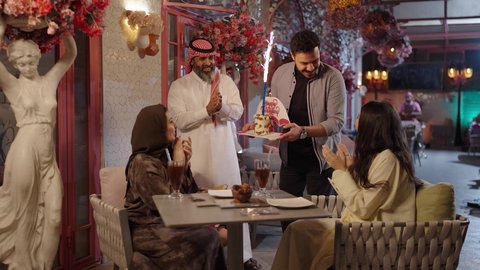 Enjoying cold drinks in the summer, enjoying recreational activities, a veiled Saudi Arabian woman wearing an abaya sits in a high-end coffee shop using her mobile phone, gestures of happiness and pleasure, spending enjoyable time outdoors