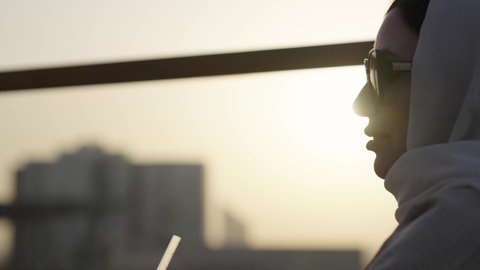 التحدث والمناقشة في كافيه راقي ، ايماءات السعادة والسرور، الوقوف بثقة والنظر إلى الكاميرا، خلفية أبراج وناطحات سحاب مدينة الرياض، الواجهة السياحية المشهورة في العاصمة، امرأة عربية خليجية سعودية محجبة ترتدي العباءة مبتسمة، الفخر والاعتزاز بالدولة السعودية.