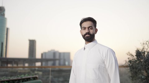 تقاطع ايدي وجدية اثناء الوقوف ، الفخر والاعتزاز بالدولة السعودية، الأبراج وناطحات السحاب المالية الإقتصادية في الرياض في الغروب، النمو والتطور العمراني في المملكة العربية السعودية، تصوير من الخلف لرجل عربي خليجي سعودي يرتدي الثوب التقليدي الابيض