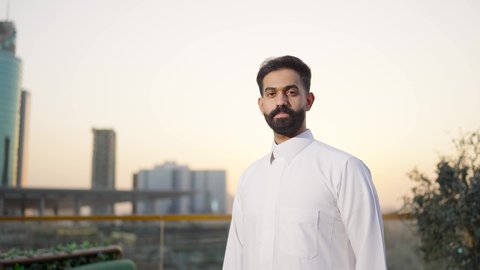 Expressions of seriousness and joy, pride and honor in the Saudi state, the towers and skyscrapers of the financial economy in Riyadh at sunset, the growth and urban development in the Kingdom of Saudi Arabia, a rear view of a Saudi Arabian Gulf man wearing the traditional white thobe.