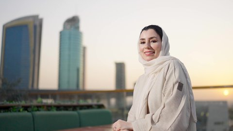 مفهوم الانوثة والجمال، ايماءات السعادة والسرور، الوقوف بثقة والنظر إلى الكاميرا، خلفية أبراج وناطحات سحاب مدينة الرياض، الواجهة السياحية المشهورة في العاصمة، امرأة عربية خليجية سعودية محجبة ترتدي العباءة مبتسمة، الفخر والاعتزاز بالدولة السعودية.