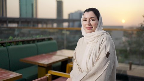 استغلال وقت الاستراحة للذهاب الى الكافيه، ايماءات السعادة والسرور، الوقوف بثقة والنظر إلى الكاميرا، خلفية أبراج وناطحات سحاب مدينة الرياض، امرأة عربية خليجية سعودية محجبة ترتدي العباءة مبتسمة، الفخر والاعتزاز بالدولة السعودية.