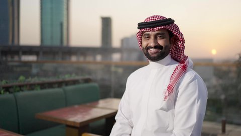 Standing in one of the upscale cafes, pride and honor in the Saudi state, the towers and skyscrapers of the financial economy in Riyadh at sunset, the growth and urban development in the Kingdom of Saudi Arabia, a shot from behind of a Saudi Gulf Arab man wearing the traditional thobe and shemagh.