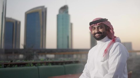 الوقوف بثقة واعتزاز بالنفس ، الفخر والاعتزاز بالدولة السعودية، الأبراج وناطحات السحاب المالية الإقتصادية في الرياض في الغروب، النمو والتطور العمراني في المملكة العربية السعودية، تصوير من الخلف لرجل عربي خليجي سعودي يرتديالشماغ والثوب التقليدي