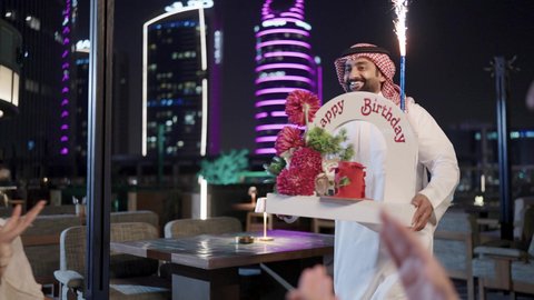 زينة الأعياد والمناسبات السعيدة، إيماءات السعادة والمفاجأة والفرح، رجل عربى خليجي سعودي يرتدي الثوب التقليدي والشماغ يقوم بالاحتفال بعيد ميلاد زوجته في إحدى المطاعم السعودية برفقة الاصدقاء، قضاء وقت ممتع مع الأهل والأصدقاء، كعكة عيد ميلاد