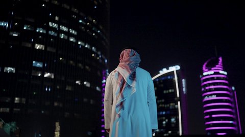 التقدم الحضاري لمدينة الرياض، الفخر والاعتزاز بالدولة السعودية، الأبراج وناطحات السحاب المالية الإقتصادية في الرياض المضيئة في الليل، النمو والتطور العمراني في المملكة العربية السعودية، تصوير من الخلف لرجل عربي خليجي سعودي يرتدي الشماغ والثوب التقليدي