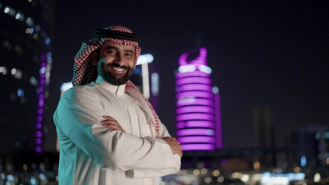 الوقوف بايدي متقاطعة، الفخر والاعتزاز بالدولة السعودية، الأبراج وناطحات السحاب المالية الإقتصادية في الرياض المضيئة في الليل، النمو والتطور العمراني في المملكة العربية السعودية، رجل عربي خليجي سعودي يرتديالشماغ والثوب التقليدي بإيماءات وجه تدل على السعادة والفرح