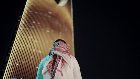 التطور والتقدم المعماري في دول الخليج،  الأبراج وناطحات السحاب المالية الإقتصادية في الرياض المضيئة في الليل، النمو والتطور العمراني في المملكة العربية السعودية، تصوير من الأسفل لرجل عربي خليجي سعودي يرتدي الثوب التقليدي بإيماءات وجه تدل على السعادة والفرح