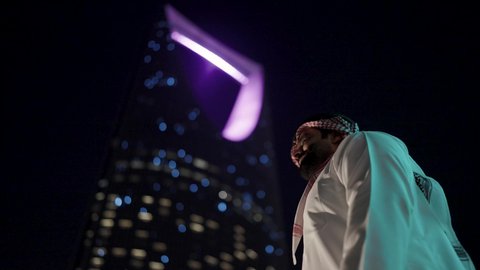 الاهتمام بالمظهر الخارجي ، الأبراج وناطحات السحاب المالية الإقتصادية في الرياض المضيئة في الليل، النمو والتطور العمراني في المملكة العربية السعودية، تصوير من الأسفل لرجل عربي خليجي سعودي يرتدي الثوب التقليدي بإيماءات وجه تدل على السعادة والفرح، مفهوم الرجولة والأناقة