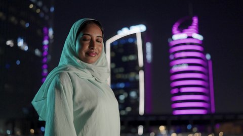 ايماءات السعادة والسرور، الوقوف بثقة والنظر إلى الكاميرا، خلفية أبراج وناطحات سحاب مدينة الرياض، امرأة عربية خليجية سعودية محجبة ترتدي العباءة مبتسمة، التقدم والتطور المعماري في دول الخليج، الفخر والاعتزاز بالدولة السعودية.