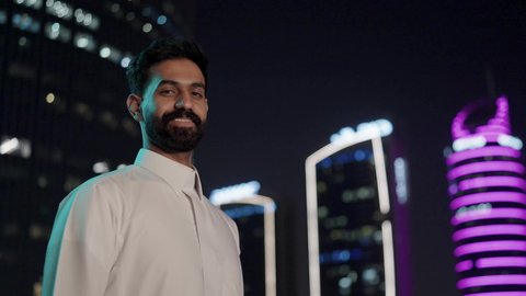 The towers and financial skyscrapers in Riyadh illuminated at night, the growth and urban development in the Kingdom of Saudi Arabia, an Arab Gulf Saudi man wearing traditional attire with facial expressions indicating happiness and joy, the concept of masculinity and elegance, the attention to external appearance.