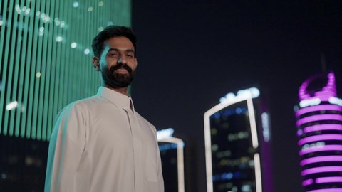 النمو والتطور العمراني في المملكة العربية السعودية، رجل عربي خليجي سعودي يرتدي الثوب التقليدي بإيماءات وجه تدل على السعادة والفرح، الأبراج وناطحات السحاب المالية الإقتصادية في الرياض المضيئة في الليل، مفهوم الرجولة والأناقة، الاهتمام بالمظهر الخارجي