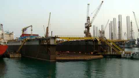 A time-lapse from day to night of the dry dock movement in Bahrain, the ambient lighting around the ships and cranes, an active industrial environment, the dynamics of continuous work.