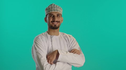الوقوف باسقامة و أيدي متقاطعة, مفهوم الأناقة و الرجولة,  الاهتمام بالمظهر الخارجي, بورتريه لرجل عربي خليجي عماني يرتدي الدشداشه والكمه ينظر إلى الكاميرا بإيماءات السعادة و السرور, التقاط صور شخصية للوثائق الرسمية،  خلفية زرقاء