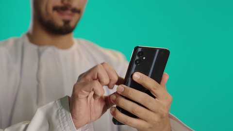 Searching for the location of something, watching enjoyable movies and series, following social media sites via mobile phone, communicating with family and friends, using technology and modern techniques, a photo of an Arab Gulf Omani man wearing a dishdasha and kumma holding a mobile device in his hand, blue background.