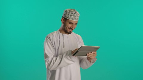 تصفح التطبيقات والمواقع، الالعاب الالكترونية على الاجهزة الحديثة، انجاز مهام العمل عن بعد، الدردشة والتواصل مع الاصدقاء، اجراء مكالمة الفيديو باستخدام جهاز تقني حديث، رجل عربي خليجي عماني مبتسم يرتدي الكمة والدشداشة يستخدم جهاز التابلت، خلفية زرقاء