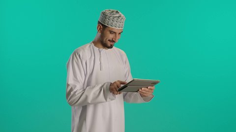 تعابير وايماءات الاندماج، الالعاب الالكترونية على الاجهزة الحديثة، انجاز مهام العمل عن بعد، الدردشة والتواصل مع الاصدقاء، اجراء مكالمة الفيديو باستخدام جهاز تقني حديث، رجل عربي خليجي عماني مبتسم يرتدي الكمة والدشداشة يستخدم جهاز التابلت، خلفية زرقاء