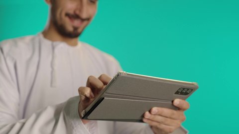 مفهوم ممارسه الانشطه والعمل عن بعد، الالعاب الالكترونية على الاجهزة الحديثة، انجاز مهام العمل عن بعد، الدردشة والتواصل مع الاصدقاء، اجراء مكالمة الفيديو باستخدام جهاز تقني حديث، رجل عربي خليجي عماني مبتسم يرتدي الكمة والدشداشة يستخدم جهاز التابلت، خلفية زرقاء