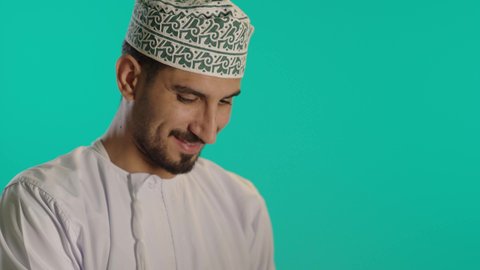 ايماءات السعادة والسرور بتحقيق الأهداف، إدارة شؤون الأعمال وتنظيمها عبر الاجهزة التقنية، دمج العمل مع التقنية والتكنولوجيا، مهن ووظائف ادارية مكتبية, تصوير لرجل عربي خليجي عماني يرتدي الدشداشة والكمة  يحمل بيده جهاز الحاسوب المحمول ، خلفية زرقاء