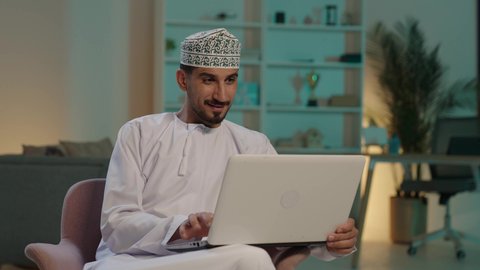 دمج العمل مع التقنية والتكنولوجيا، مهن ووظائف ادارية مكتبية, إدارة شؤون الأعمال وتنظيمها عبر الاجهزة التقنية، ايماءات السعادة والسرور بتحقيق الأهداف، تصوير  لرجل عربي خليجي عماني يرتدي الدشداشة والكمة  يحمل بيده جهاز الحاسوب المحمول