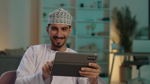 Electronic games on modern devices, completing remote work tasks, chatting and communicating with friends, making video calls using modern technology, a smiling Arab Gulf Omani man wearing a kumma and dishdasha using a tablet, the concept of practicing activities and remote work.